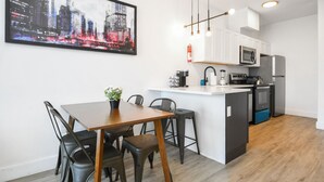 A modern kitchen with white cabinets, a black refrigerator, and a microwave. There's a dining area with a wooden table and chairs, and a cityscape painting on the wall. - Beautiful Studio Near EWR bahz (Elizabeth)
