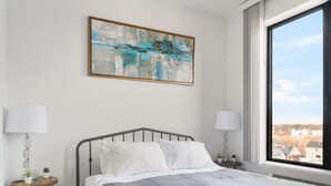 A modern bedroom with a metal bed frame, white bedding, a blue striped rug, a cityscape view through a large window, and a painting on the wall. - Beautiful Studio Near EWR bahz (Elizabeth)