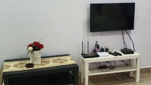 A room with a white TV on the wall, a small table with a vase of flowers, and a white shelf with electronic devices. - OYO Home 90496 Delima Homestay (Alor Setar)