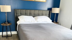 Un dormitorio con una cama tapizada de gris, dos mesitas de noche con lámparas, y un cuadro abstracto de gran tamaño en la pared.