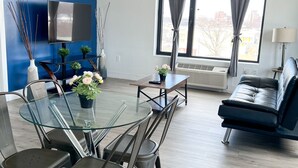 Una sala de estar moderna con una mesa de comedor de cristal, sillas oscuras, un sofá azul, un televisor de pantalla plana y un ventanal con cortinas.