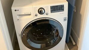 Una lavadora LG Direct Drive compacta con panel de control, una plancha pequeña y una tabla de surf apoyada en la pared.