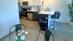Una cocina moderna con armarios blancos, un microondas y un fregadero. Hay una zona de comedor con una mesa y sillas de vidrio, y una planta en maceta.