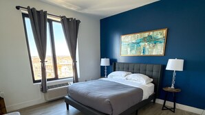Un dormitorio con cama, dos mesitas de noche, una ventana con cortinas, un cuadro en la pared y vistas de la ciudad.