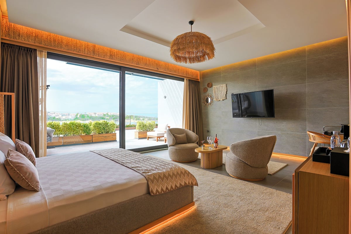 Una habitación de hotel moderna con una cama grande, televisión, balcón con vistas y un pequeño rincón de descanso.
