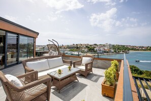 A balcony with wicker furniture, a glass table, and a view of the sea and town. - Cesme Sarezya Boutique Hotel  (Cesme)