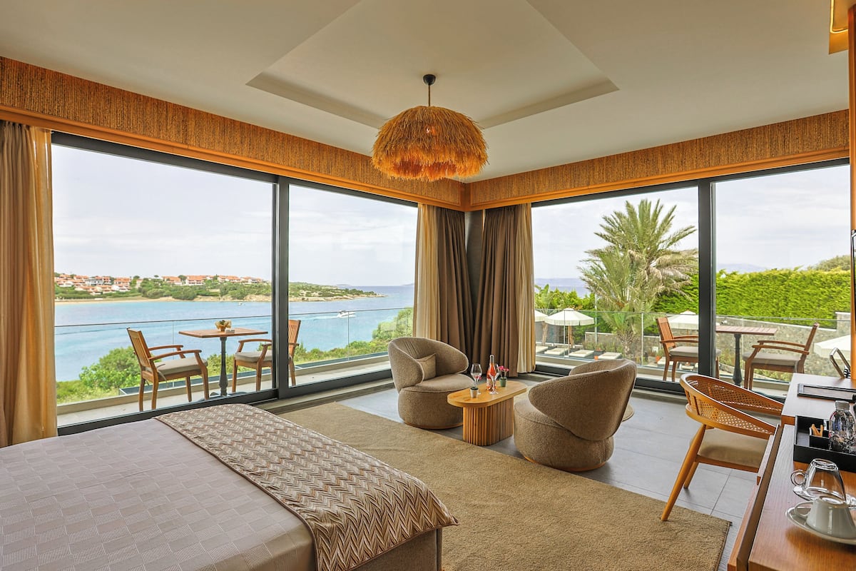 Una habitación de hotel moderna con una cama grande, dos sillones, una mesa de centro y un balcón con vistas al mar y al verdor.