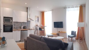 A modern living room with a grey sofa, a dining area with a small table and chairs, a kitchen with white cabinets and a microwave, and a TV mounted on the wall.