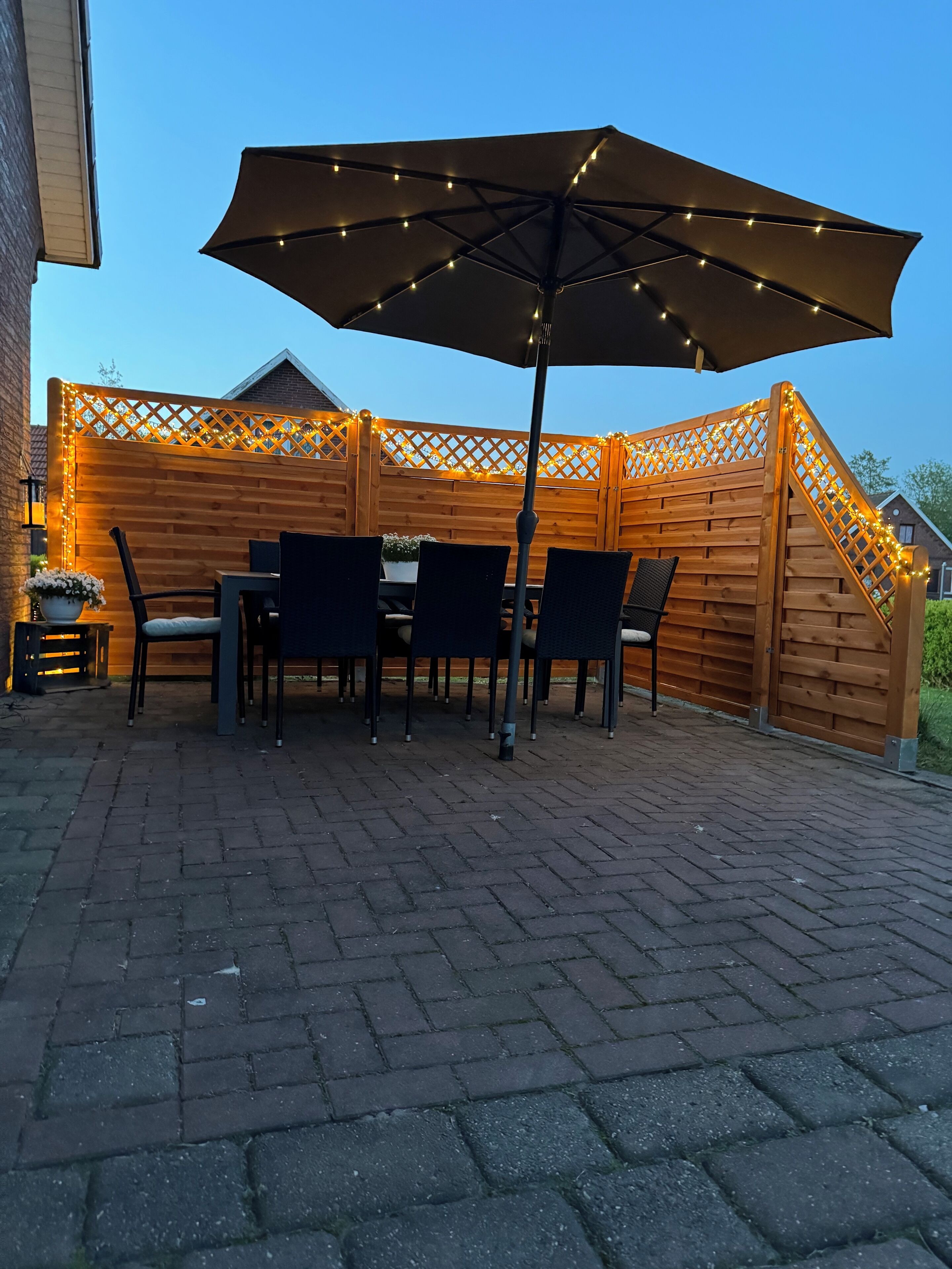 Een terras met een houten tafel en stoelen, een parasol en lichtjes.