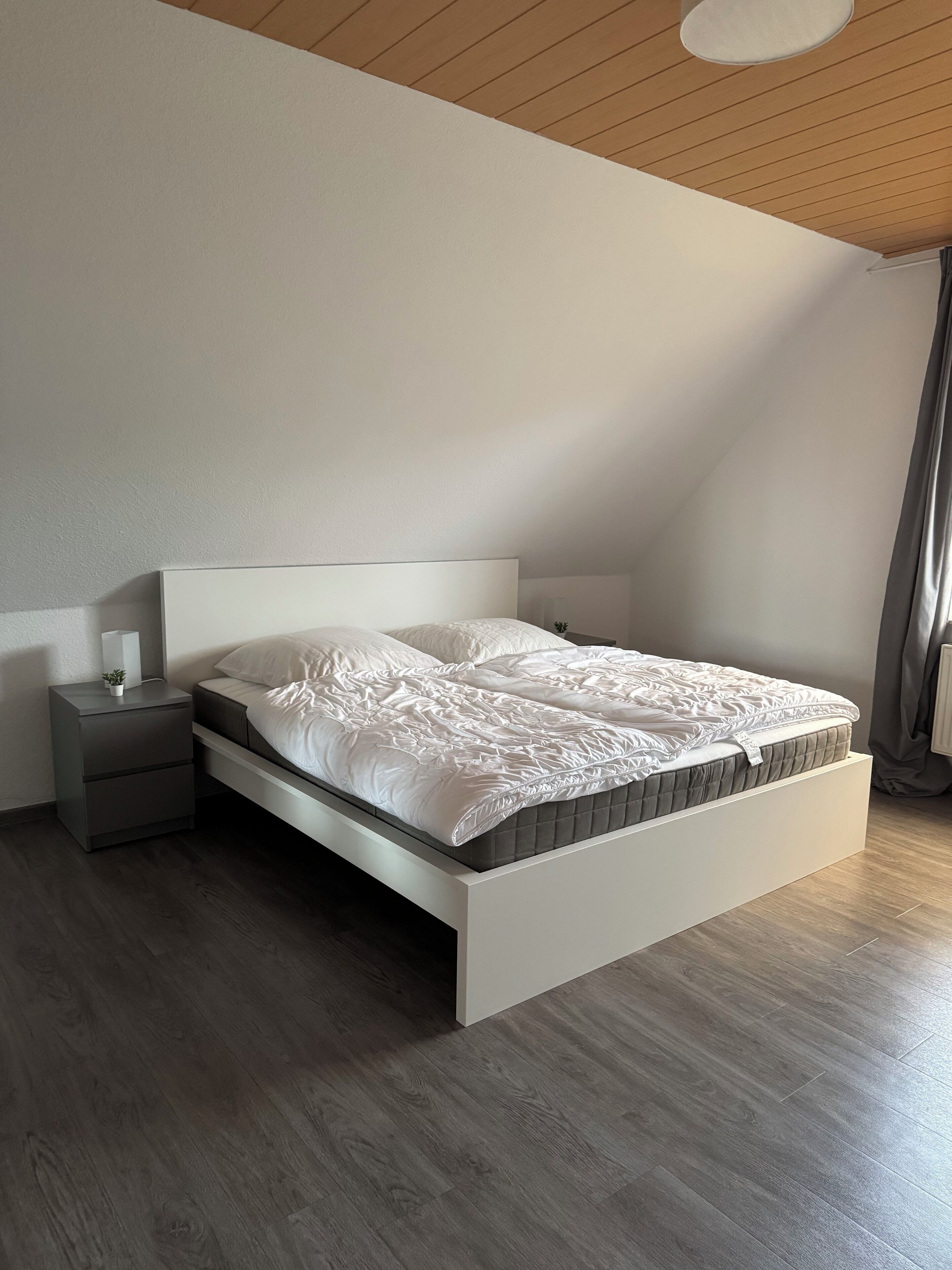 Un dormitorio moderno con una cama blanca, una mesita de noche gris y una pequeña planta en maceta.