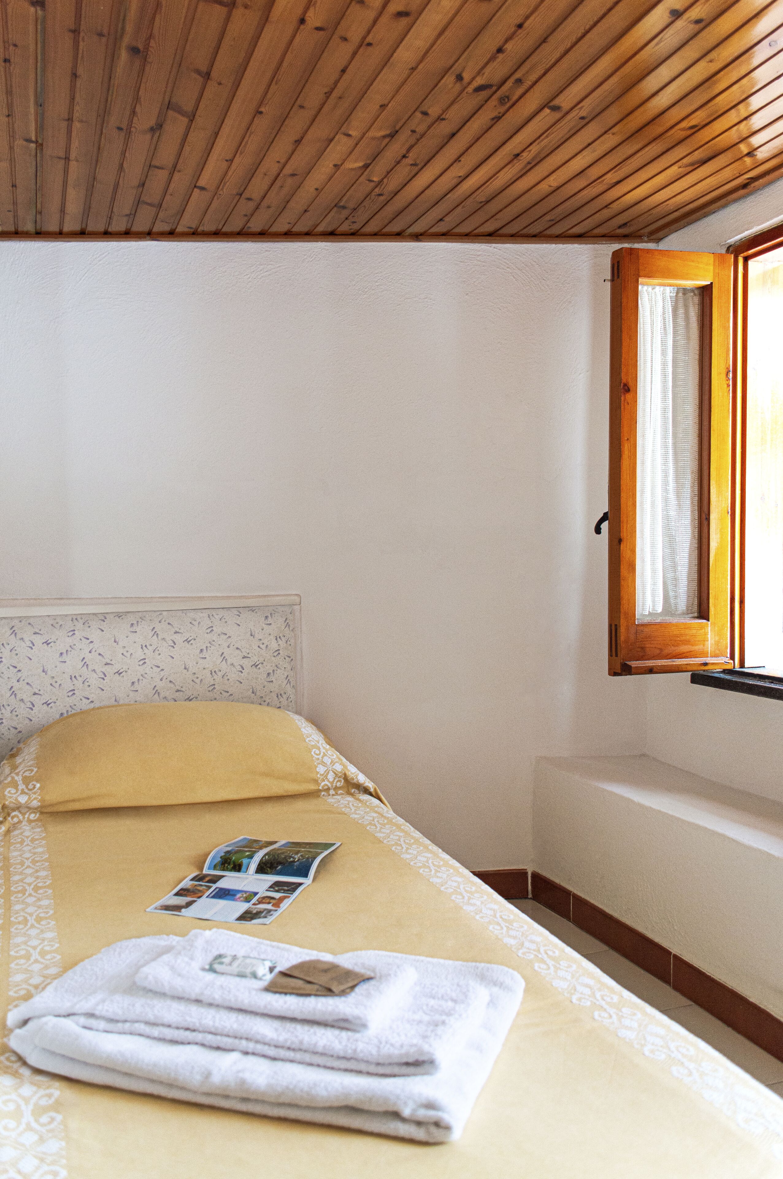 A small, single-bed room with a wooden ceiling, a window with a wooden frame, a bedside table, and a bed with a yellow pillow and a folded towel.