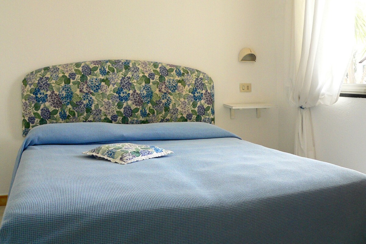A bed with a floral headboard, a blue bedspread, and a pillow. There is a small white shelf on the wall and a window with curtains.