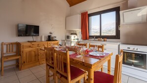 A dining area with a wooden table set for four, a television mounted on the wall, and a kitchen area with a microwave and oven. - Le Bleuet du Lac - Appartement avec Terrasse Vue Lac (Savines-le-Lac)
