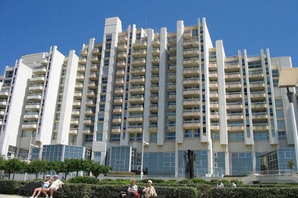 A high-rise residential building with multiple balconies, a modern architectural design, and a landscaped area with benches.
