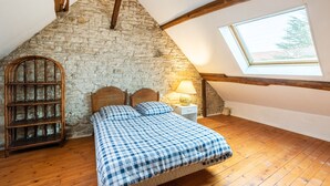 Une chambre avec un mur en pierre, des poutres en bois, un plafond en pente, un lit recouvert d’une courtepointe à carreaux, une table de chevet avec une lampe et une fenêtre donnant sur les arbres.
