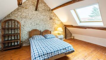Slaapkamer met een stenen muur, houten balken, een schuine kap, een bed met een geruit beddegoed, een nachtkastje met een lamp en een raam met uitzicht op bomen.
