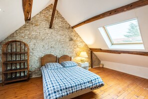 Slaapkamer met een stenen muur, houten balken, een schuine kap, een bed met een geruit beddegoed, een nachtkastje met een lamp en een raam met uitzicht op bomen.