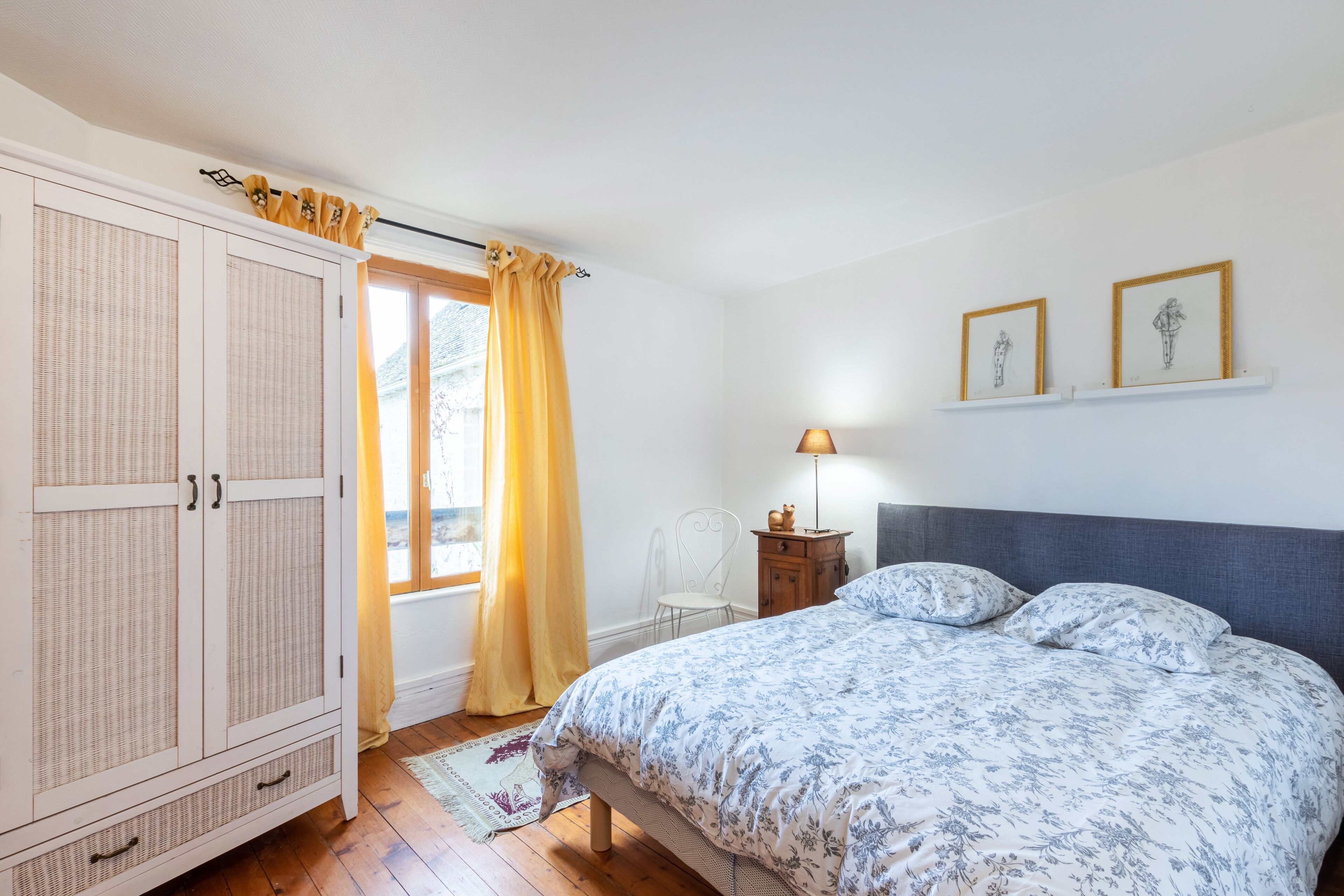 Quarto com cama, guarda-roupa, janela com cortinas, cadeira, mesa de cabeceira e duas fotografias emolduradas na parede.