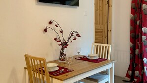 A dining area with a wooden table, two chairs, a vase with red berries, a wine glass, and a flat-screen TV mounted on the wall.