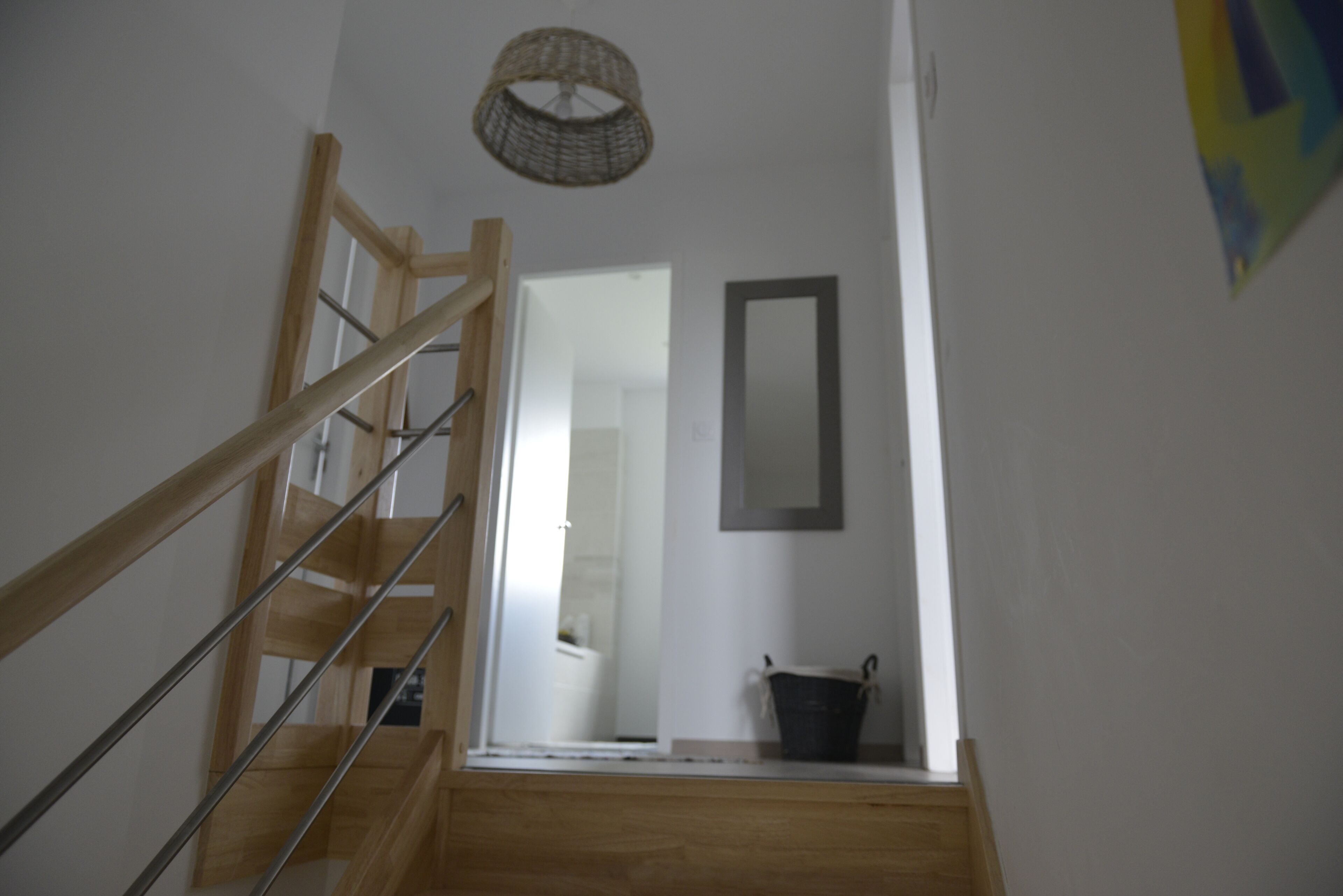 A wooden staircase with a metal handrail leading to a room with a white wall, a hanging basket, and a framed picture.
