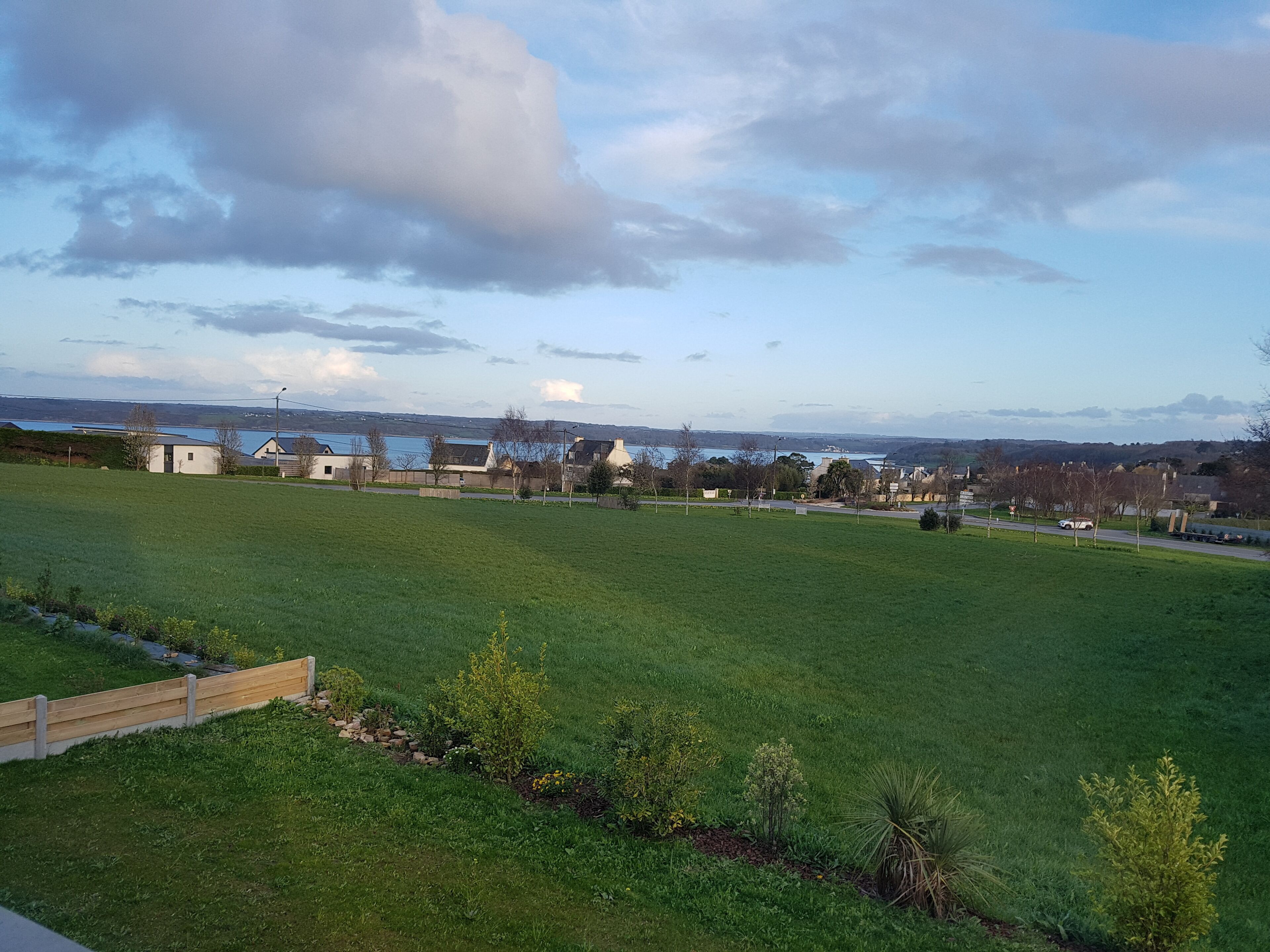 Een grasveld met een houten omheining, een weg en huizen in de verte.