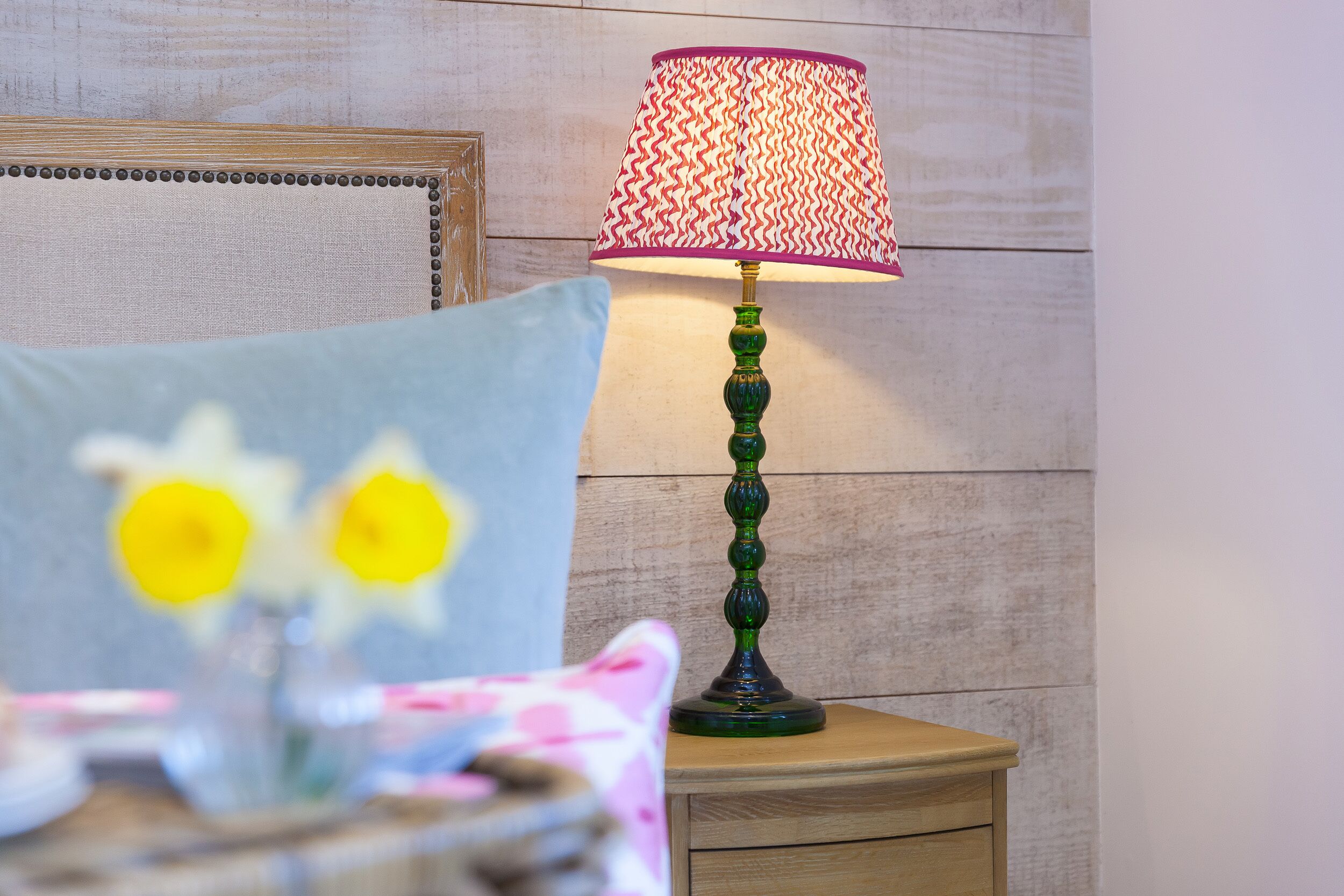 A room with a wooden wall, a green lamp with a patterned lampshade, and a blue pillow with daffodils.