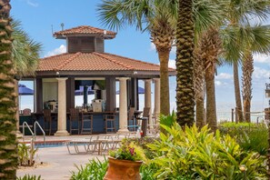 Relax by the pool surrounded by palm trees and lounge chairs, with a central bar area and covered seating.