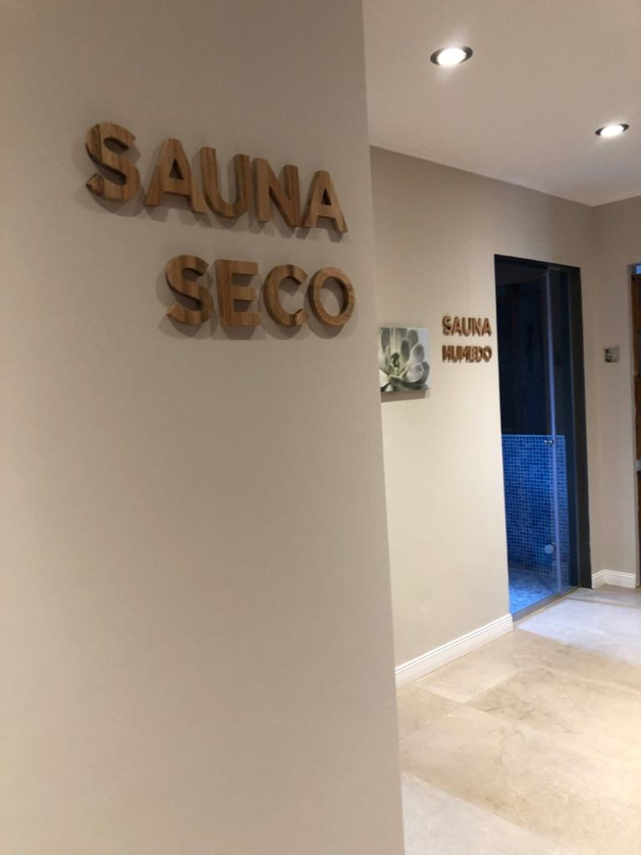 Un pasillo con adornos de madera en la pared que deletrean “SAUNA SECO” y una puerta que indica “SAUNA HUMEDO”.
