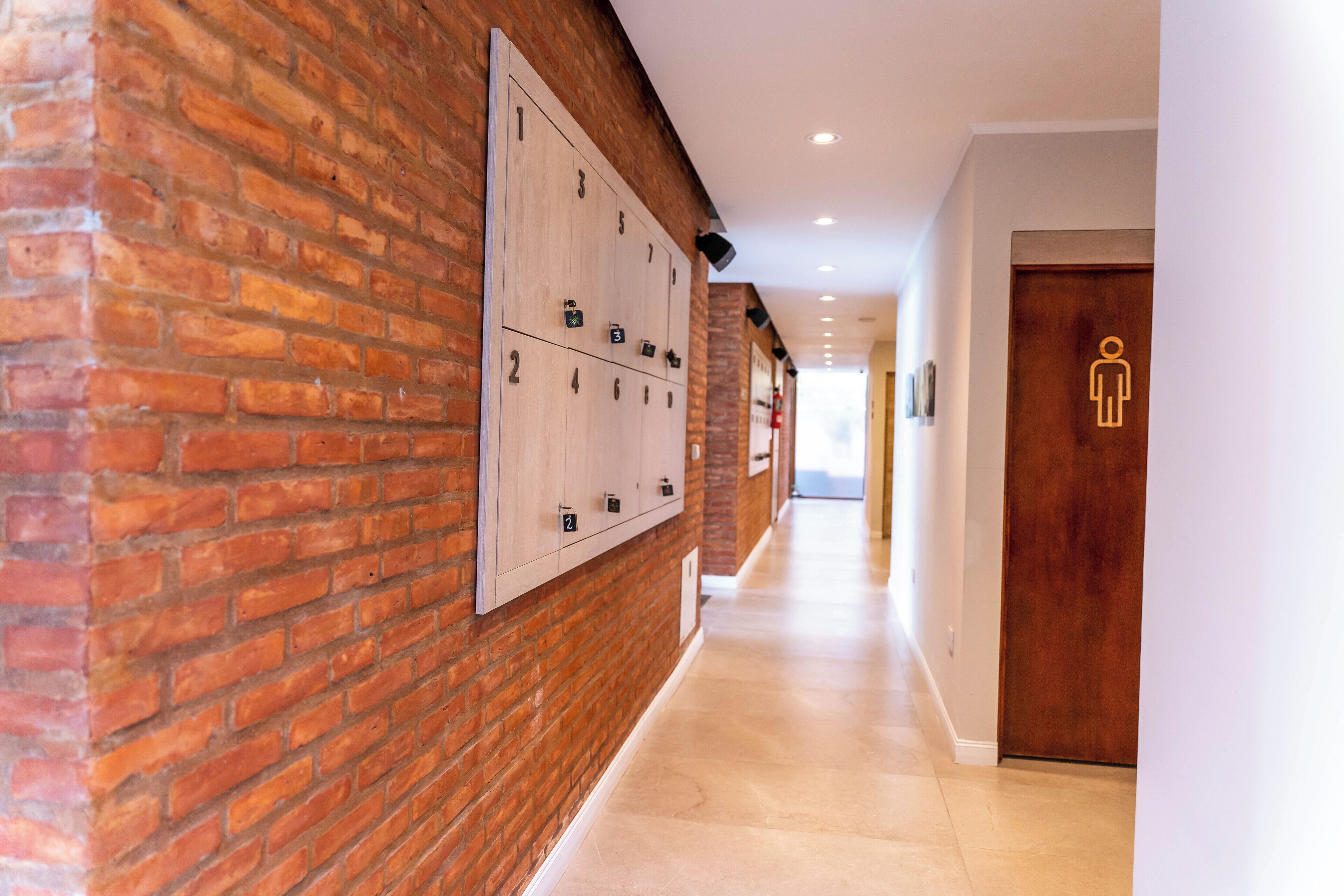A hallway with a brick wall, lockers, and a wooden door with a restroom symbol.