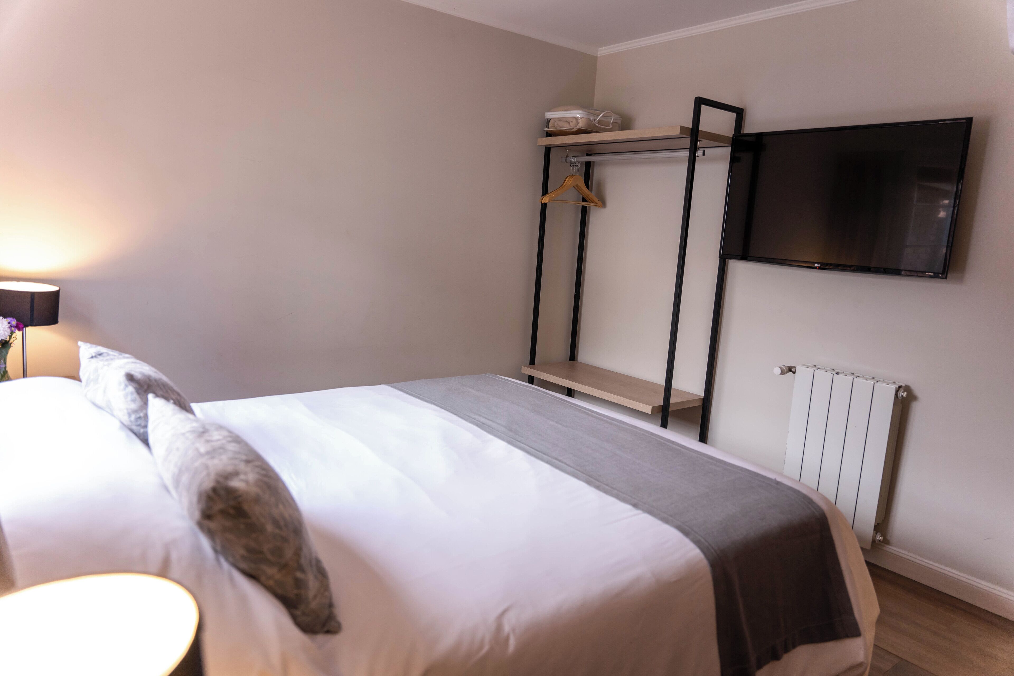 A hotel room with a bed, a TV mounted on the wall, a radiator, and a shelf with a hanging garment.