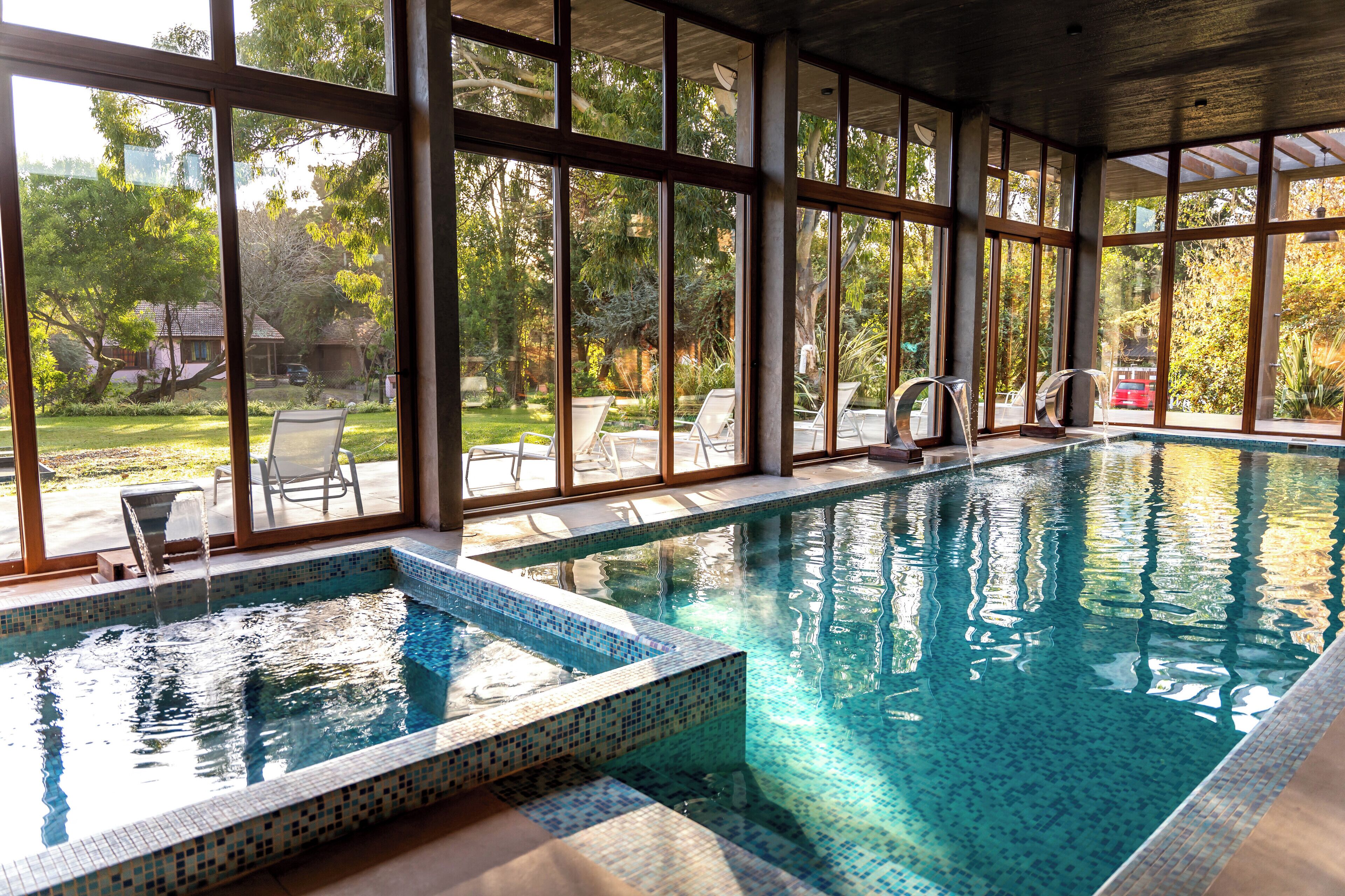 Une piscine intérieure aux parois en verre transparent, entourée de chaises longues et offrant une vue sur un jardin.