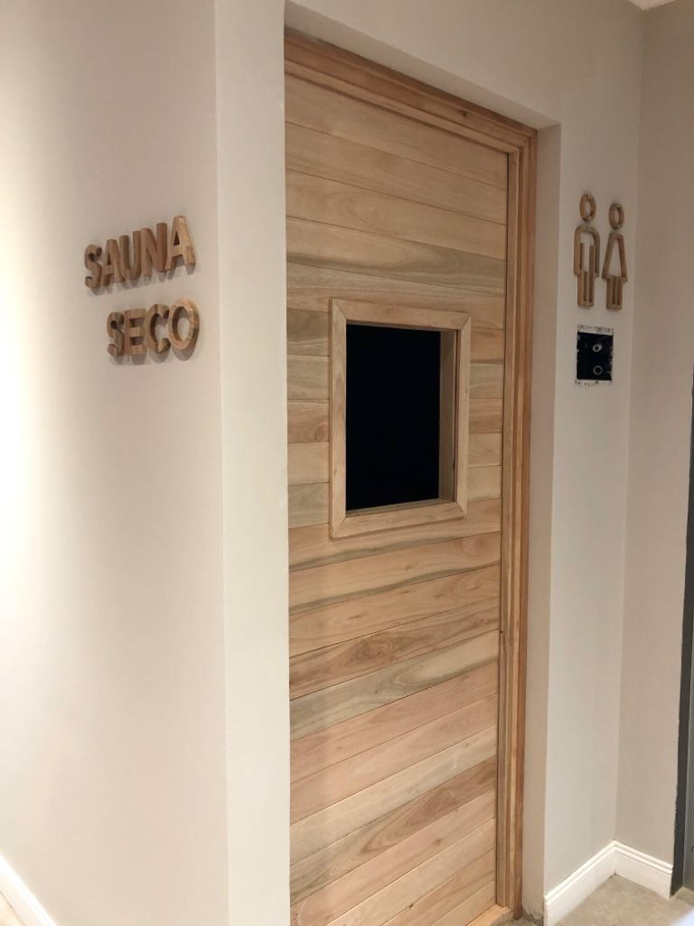 Un mur en bois avec une petite fenêtre et une porte, et une pancarte sur laquelle on peut lire « SAUNA SECO ».
