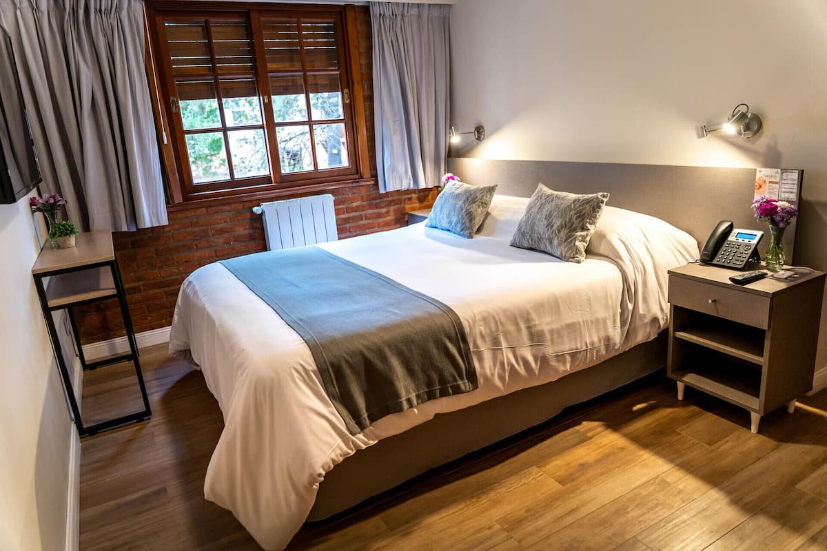 Un dormitorio ordenado con una cama grande, una mesita de noche con un teléfono y una ventana con persianas.