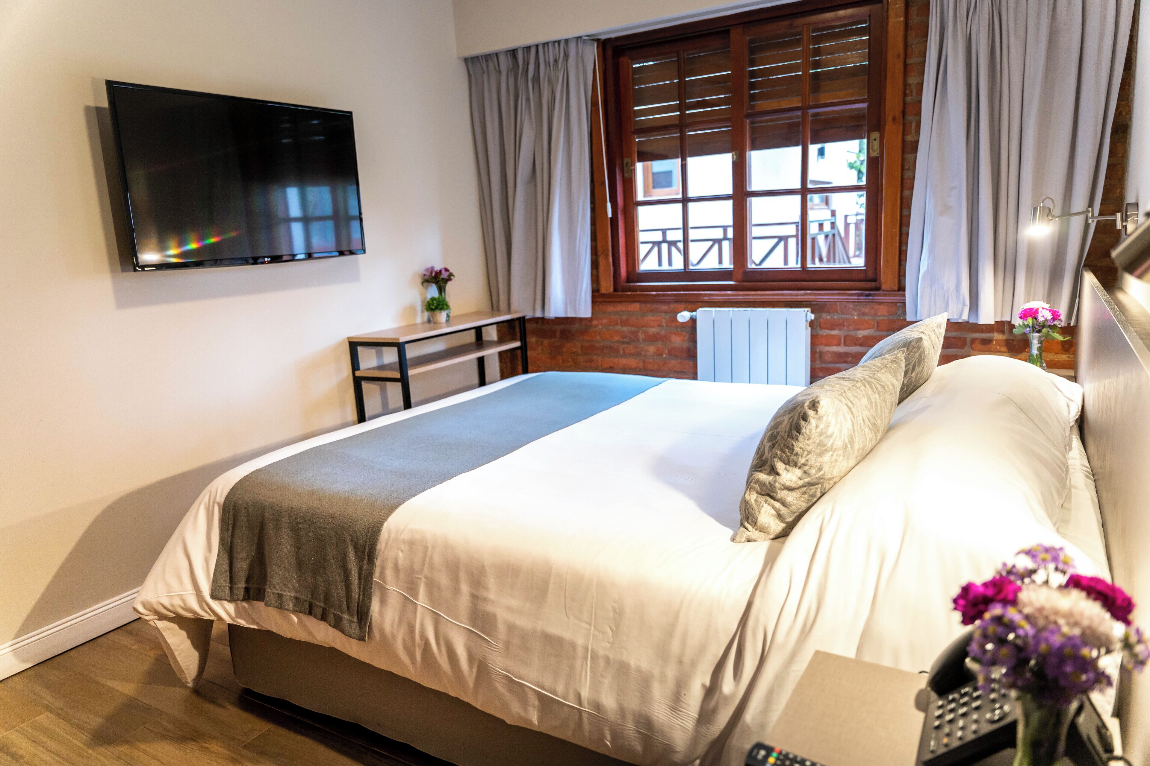 Una camera da letto con un letto, una TV fissata al muro, una finestra con tende, un tavolino con un vaso di fiori e un termosifone.