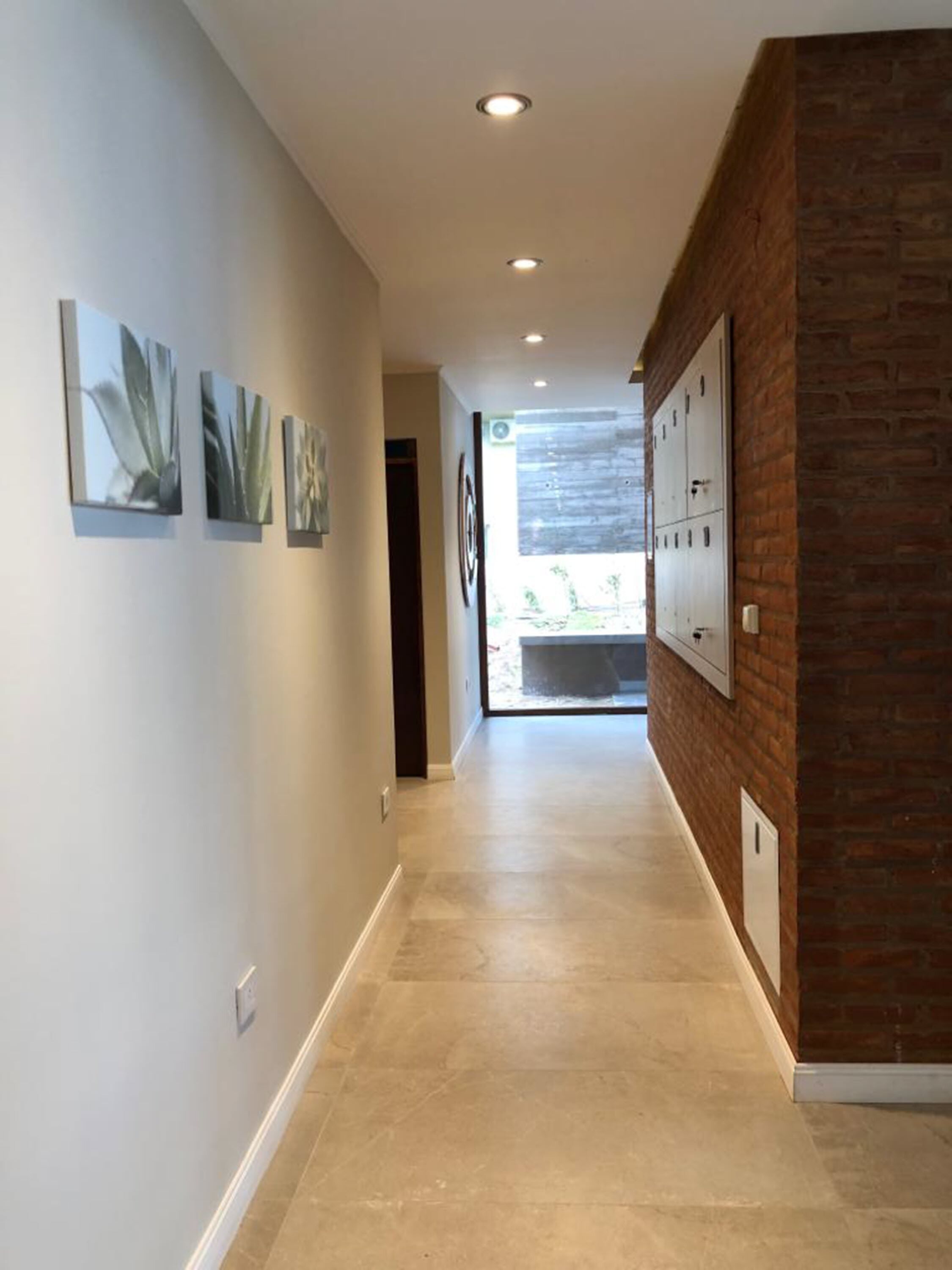 Un pasillo de diseño moderno con un muro decorativo de ladrillo, luces empotradas en el techo y vistas al exterior a través de una puerta de vidrio.