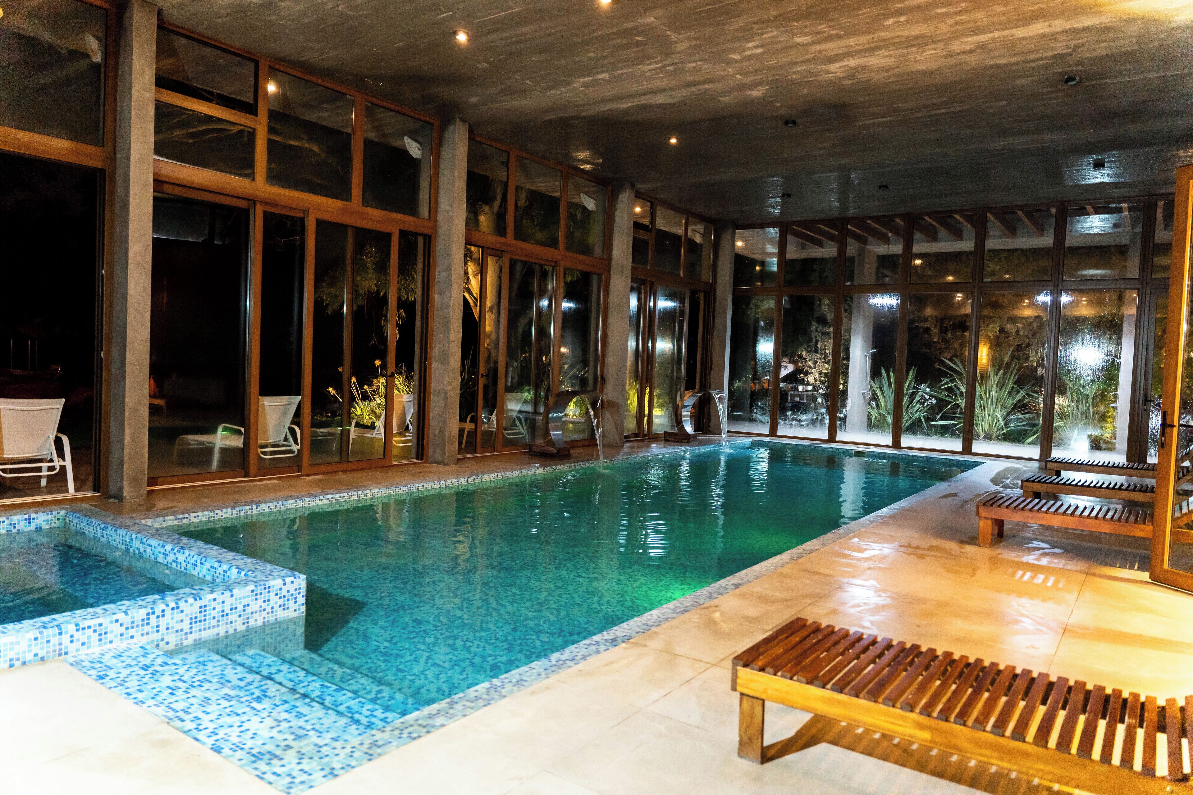 An indoor swimming pool with clear glass walls, wooden deck seating, and a tiled floor.