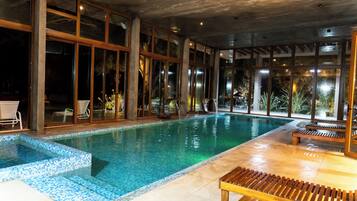 An indoor swimming pool with clear glass walls, wooden deck seating, and a tiled floor.