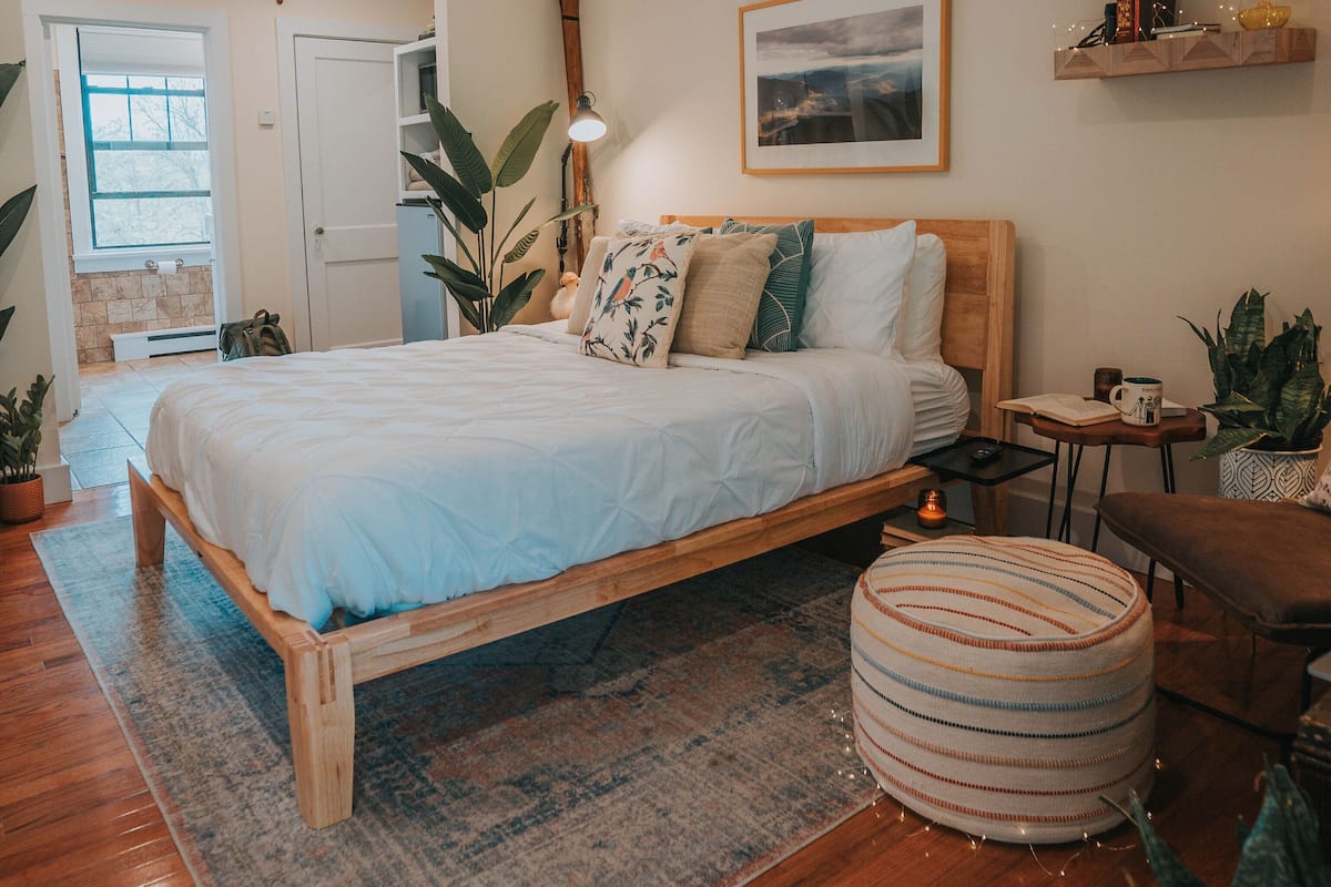 A bedroom with a wooden bed, a round ottoman, a small table with a lamp, and a window with a view of the outdoors.