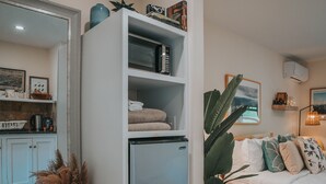 A compact living space with a built-in refrigerator, a bed with a view, a small seating area, and a kitchen area visible through an open door. - Willow Pond Motel (Catskill)