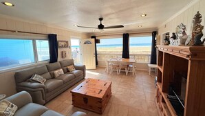 Ett vardagsrum med en soffa, ett soffbord och ett matbord, stora fönster med utsikt över stranden och en takfläkt.