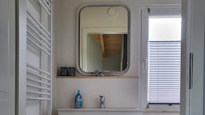 Ein Badezimmer mit weißem Waschtisch, einem Spiegel, einem Heizkörper, einem kleinen Ablagefach für Pflegeprodukte und einem Fenster mit Jalousien.