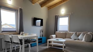 Ein Wohnzimmer mit einem weißen Sofa, einem Esstisch mit Stühlen, einem an der Wand montierten Fernseher und einem Fenster mit Vorhängen.
