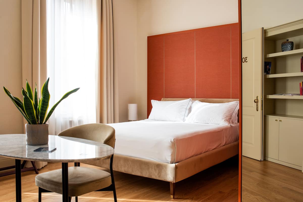 A hotel room with a bed, a round table with a chair, a potted plant, and a bookshelf.