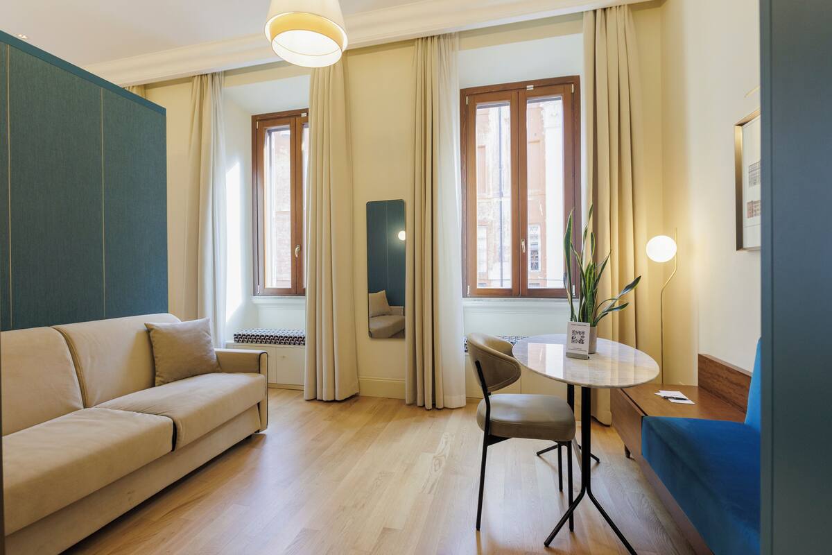 A modern living room with a sofa, a dining table with chairs, and large windows with curtains.