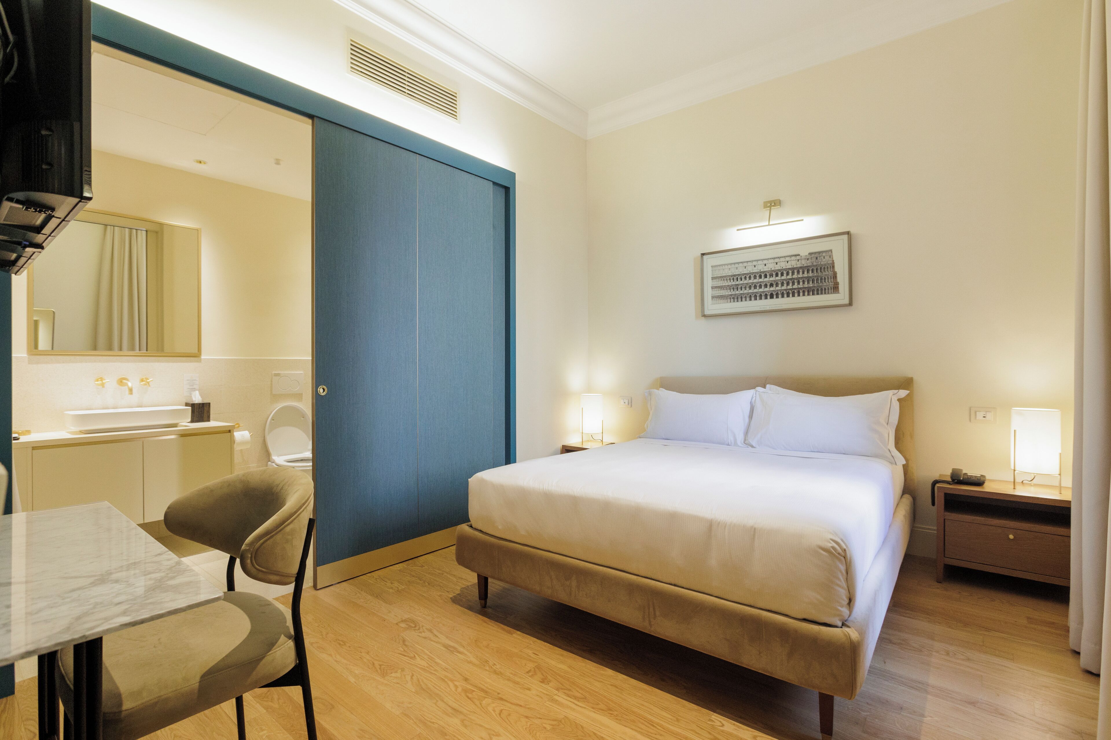Una camera d'albergo con un letto, una scrivania con una sedia, un bagno e un quadro appeso al muro.