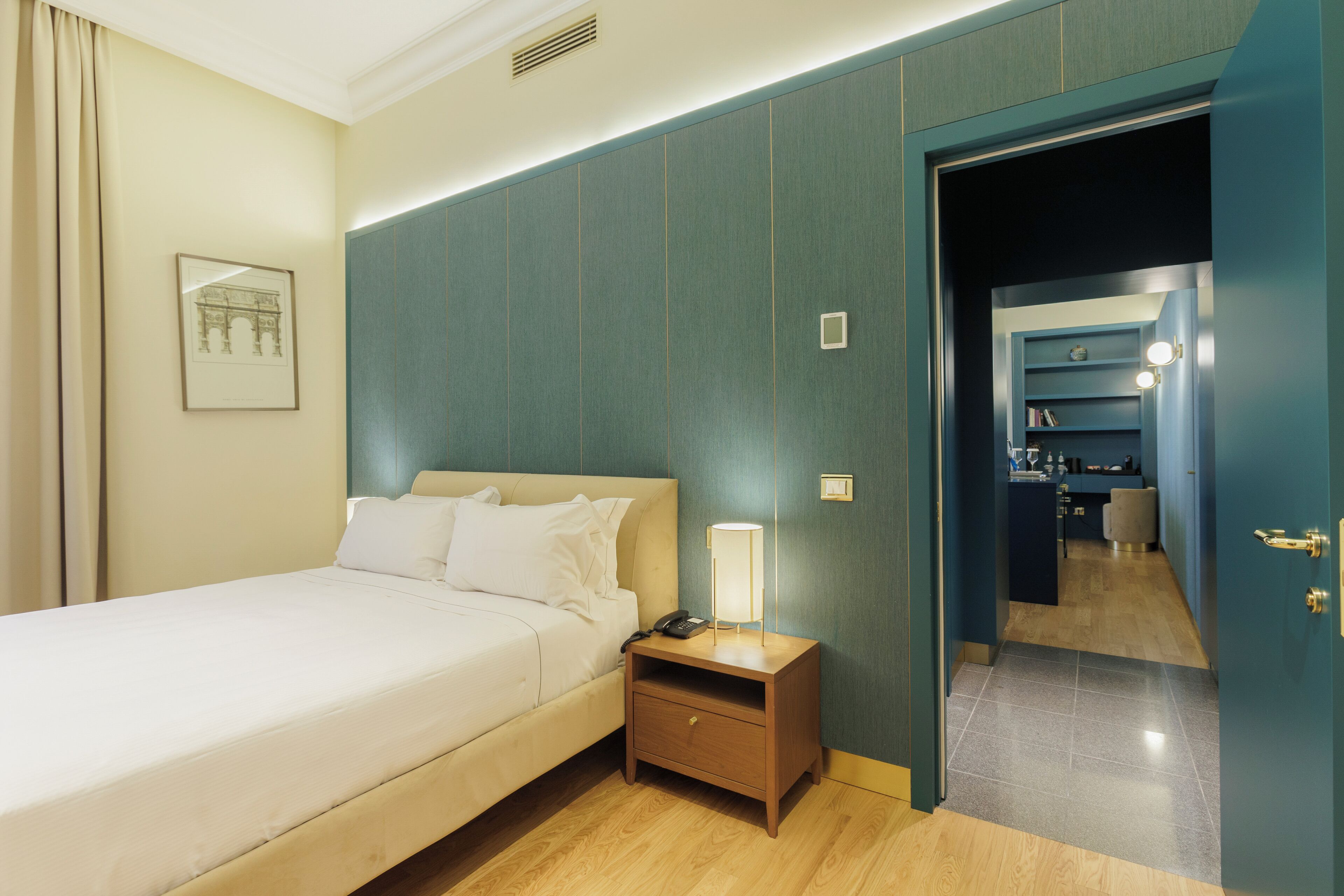 A hotel room with a bed, bedside table with a lamp, a framed picture on the wall, and a door leading to a bathroom.