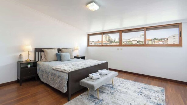 A bedroom with a large bed, a nightstand, a bench, a window with a view of buildings, and a ceiling light. - Spacious Duplex | Jacuzzi & Terrace by HostWise (Porto)