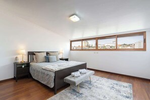 Une chambre à coucher avec un grand lit, une table de chevet, un banc, une fenêtre donnant sur des immeubles et un luminaire au plafond.