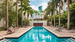 A swimming pool surrounded by palm trees and lounge chairs, with a view of tall buildings in the background. - Spacious studio apartment with a balcony (Miami)