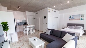 A modern, well-lit living area with a grey sofa, white coffee table, and a dining table with chairs. A kitchen is visible in the background, and there is a bed with a grey headboard in the foreground. - Spacious studio apartment with a balcony (Miami)